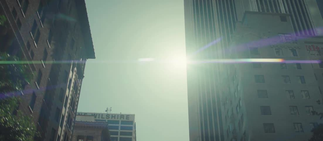 This is a lowangle shot looking up between two tall buildings in a city on a bright sunny daythe camera pans upward revealing the sky and a strong lens flare from the sun highlighting the height and scale of the surrounding architecture