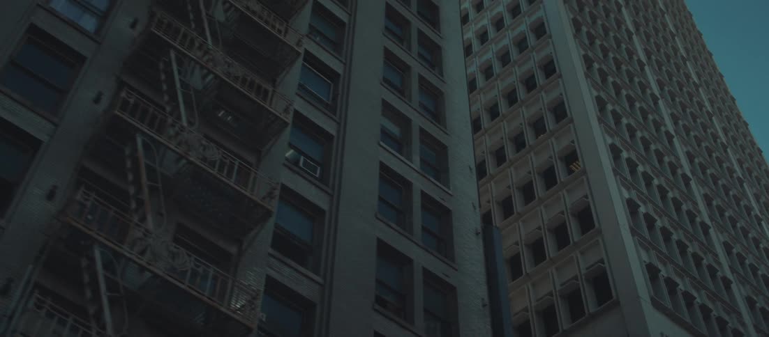 The video features a dramatic lowangle shot panning upwards to reveal the tall facades of several buildings in a city setting the architecture includes a modern tower with repetitive window patterns and an older brick building featuring several fire es
