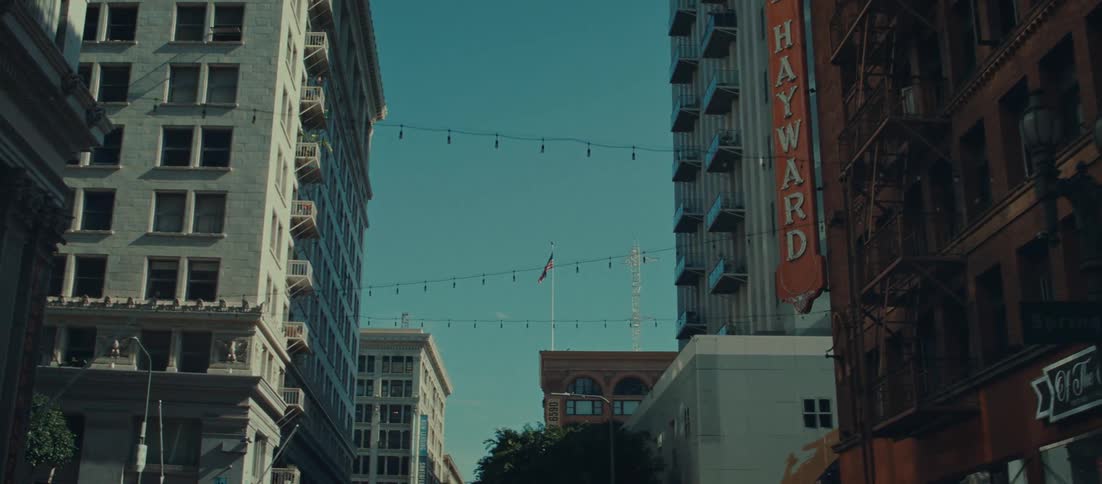 The view looks up a narrow city street framed by tall historic buildings on both sides in the center an american flag flies from a pole atop a shorter building and string lights hang across the street overhead