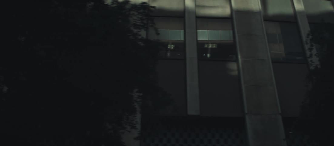 The camera quickly pans down the vertical facade of a tall modern building in an urban area passing over a sign starting with a bright sunflared view of the sky and ending on the darker lower section of the building exterior