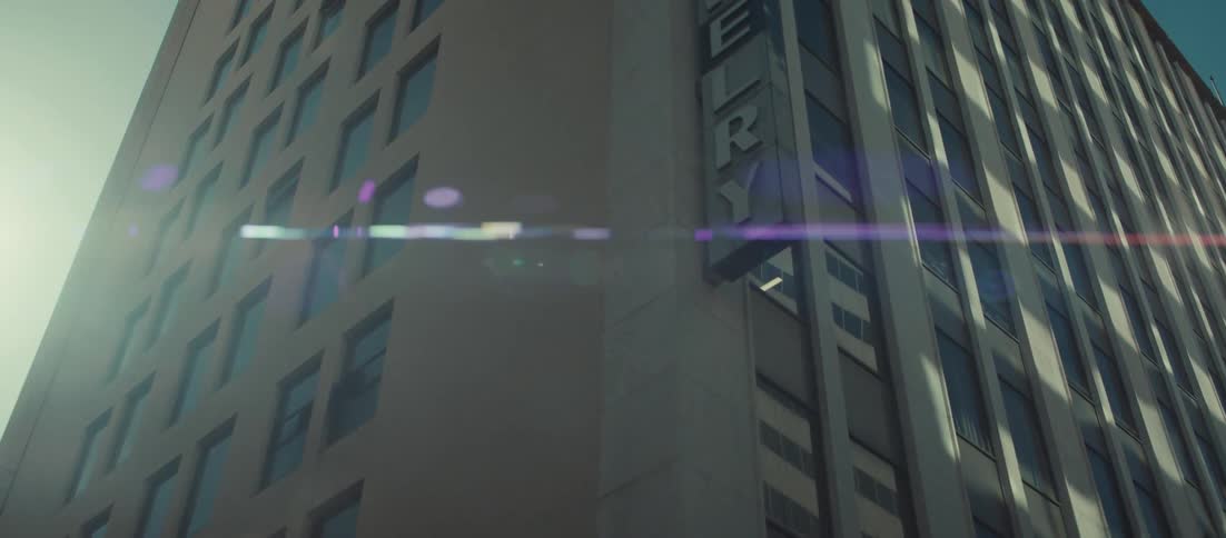 The camera tilts up the side of a tall commercial building with a grid of square windows and large vertical columns revealing a street lamp post and a sign mounted vertically on the corner of the structure