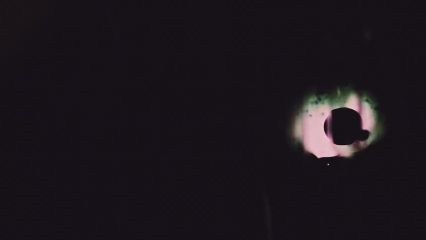 The video features an abstract dark frame where a small circular shape glowing with colorful changing light starting pinkpurple and shifting to green appears and shrinksthe dark circle within the glowing light moves and disappears behind the lig
