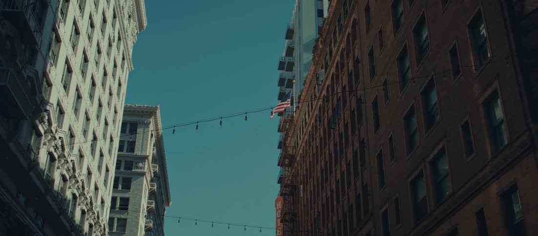 A lowangle tracking shot moves slowly between tall city buildings capturing hanging string lights and an american flag against the clear blue sky