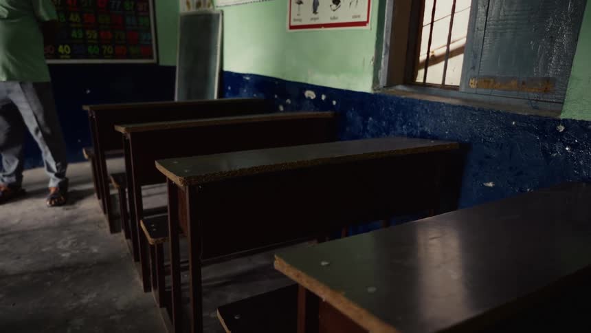 A row of simple dark wooden school desks is filmed inside a classroom with twotoned walls illuminated by natural light entering through an open window while a man in dark pants walks slowly in the far left background