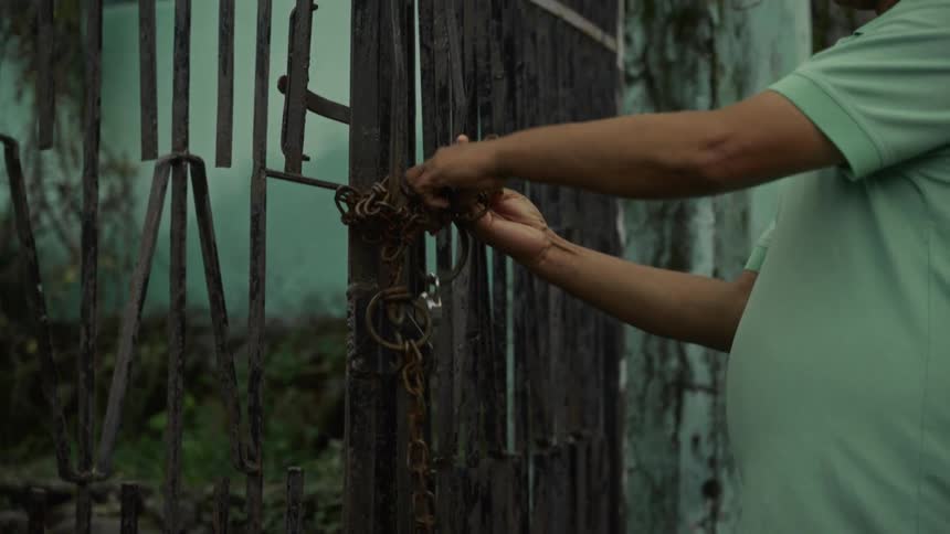 A closeup shot focuses on the hands and arms of a man in a light green shirt as he manipulate keys to unlock a padlock securing a heavy rusty metal chain wrapped around the bars of a worn dark metal gate