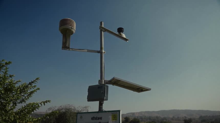 A low angle view of an outdoor weather station mounted on a pole with solar panels against a blue sky above a rural landscape