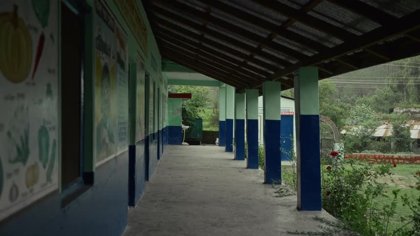 The exterior covered hallway of a simple school building is shown featuring concrete pillars painted blue and light green a concrete floor a low railing bordering a grassy yard and colorful educational posters lining the classroom wall