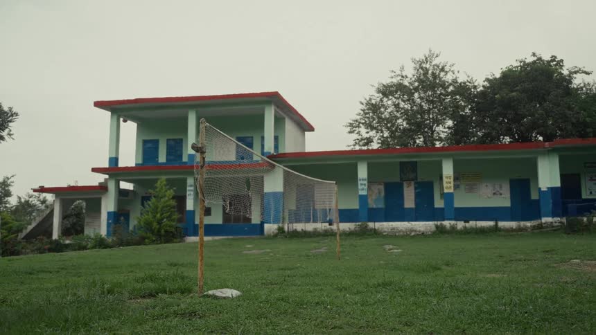 A wide shot captures a brightly painted twostory school building with a flat roof standing behind a grassy field where a loosely hung volleyball net is stretched between two makeshift poles