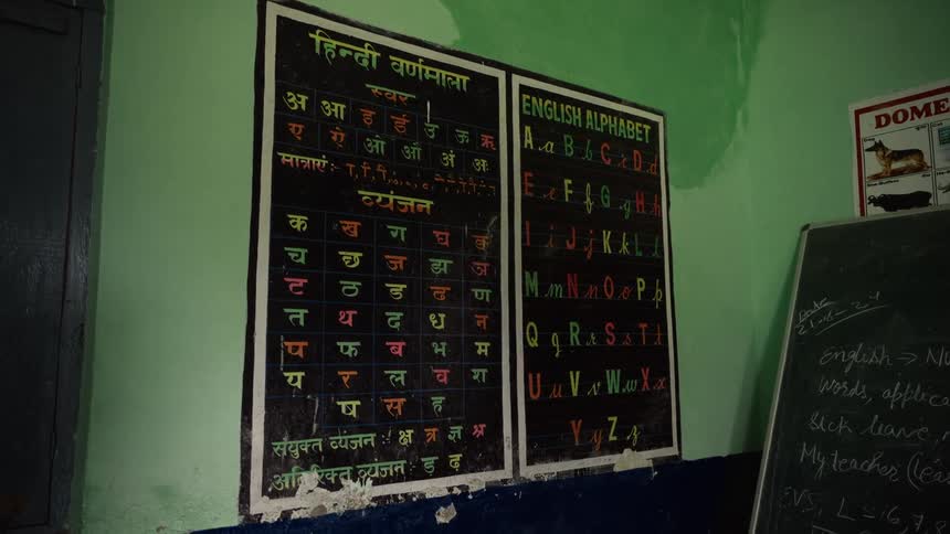 A school classroom displays hindi and english alphabet charts on a green wall beside a blackboard