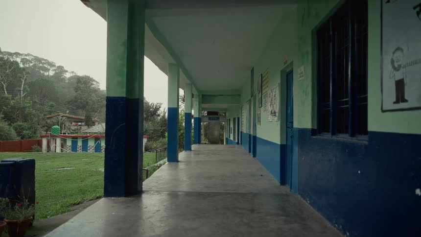 A long covered outdoor school corridor with light blue walls dark blue columns and a view of a grassy field and dense hillside