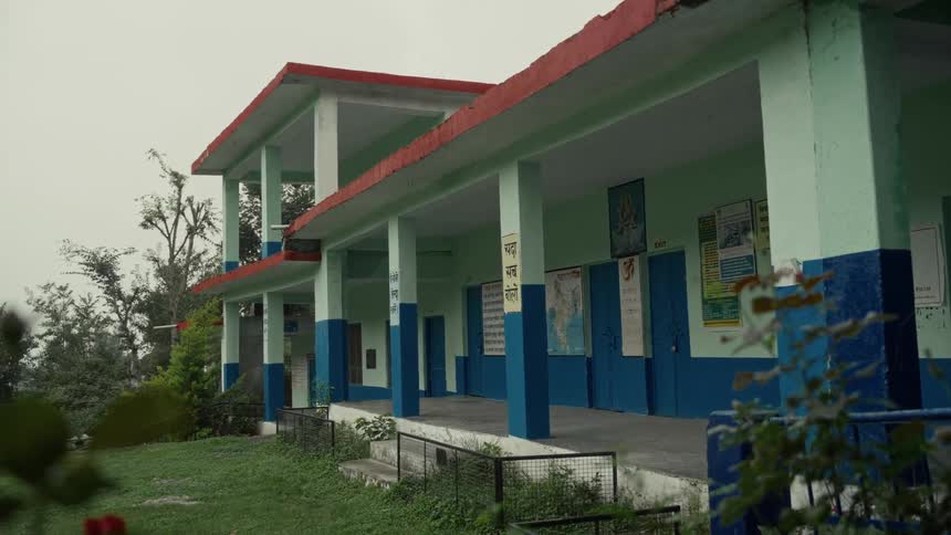 The video shows a lowangle view of a humble twostory school building with a red roof green walls and blue trim around the doors and pillars the school appears to be located in a hilly or rural area surrounded by greenery under an overcast sky