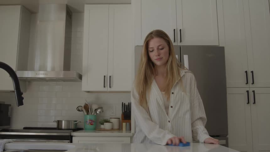A young woman with long blonde hair uses a blue sponge to wipe down a white kitchen counter in a modern kitchen with stainless steel appliances
