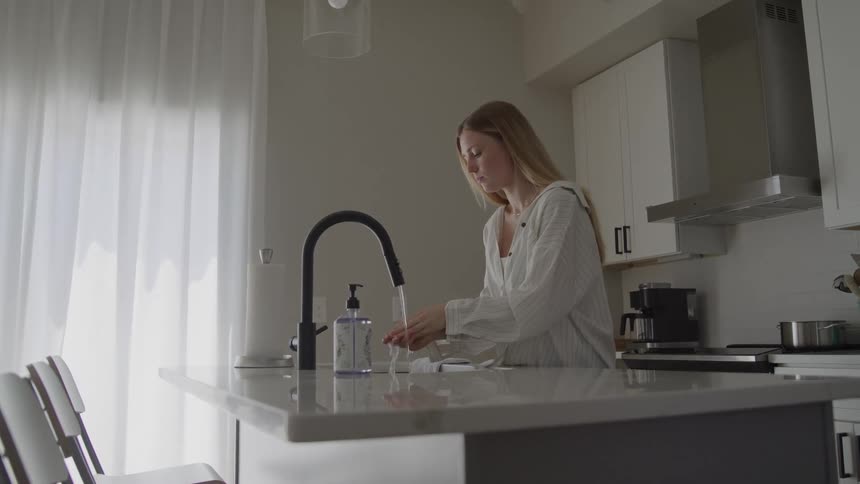 A young woman with long hair stands at the sink of a bright modern kitchen and thoroughly washes her hands using a pump of soap dispenser while running water from the high black faucet