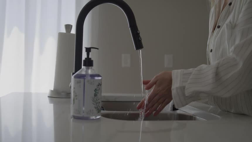 A woman wearing a white striped shirt washes her hands under a modern black faucet in a bright kitchen with a paper towel roll and soap bottle nearby