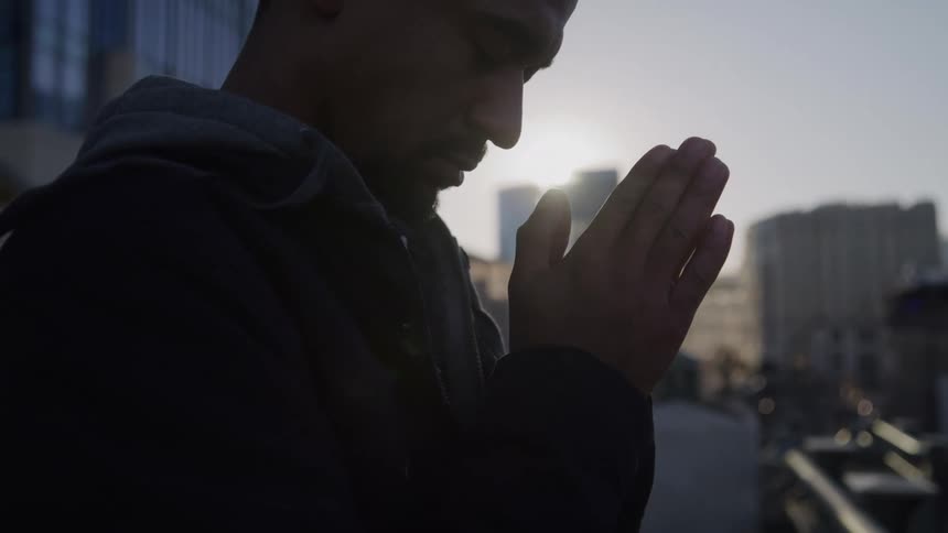 A man with a shaved head and dark jacket stands in silhouette outdoors holding his hands together in prayer or contemplation against a bright cityscape background