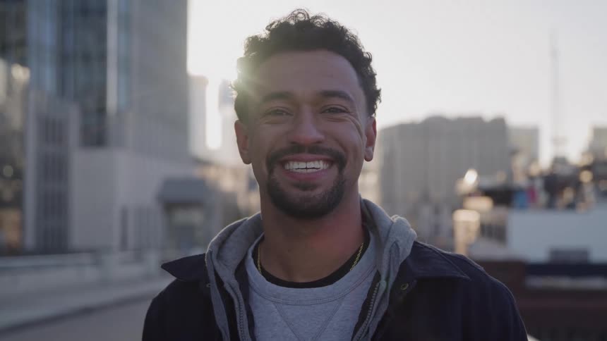 A man with curly hair and a beard is standing outdoors in an urban environment smiling directly at the camera he is wearing a jacket over a grey hooded sweatshirt and the background shows city buildings and bright slightly hazy sunlight