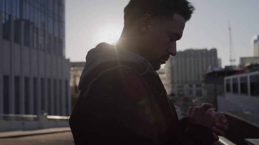 This closeup video shows a man standing on an elevated surface in a city environment looking downward with his hands clasped togetherthe sun is low and bright behind him creating a strong backlight that outlines his profile against the backdrop of