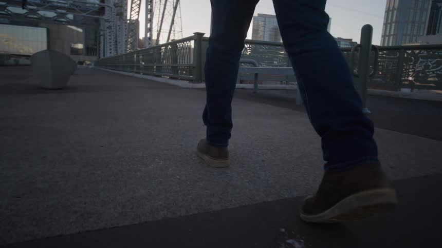A person wearing dark blue jeans and brown shoes with thick soles as he walks forward across the concrete pedestrian walkway of a large industrialstyle steel bridge with the structural framework and distant cityscape visible