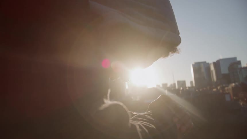A man wearing a hooded sweatshirt leans on a railing in profile on a sunny day looking down with a bright city skyline visible in the background