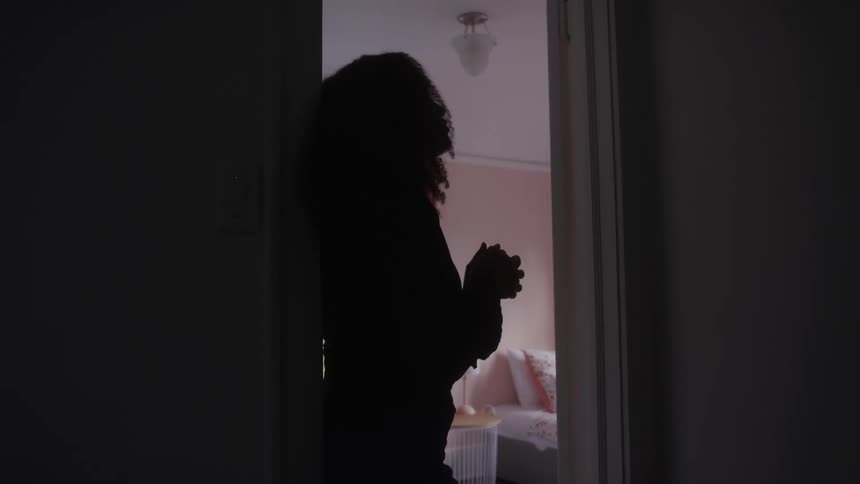 A woman stands in a dark doorway silhouetted against a softly lit room beyond her hands are held together near her chest and she appears to be looking upwards