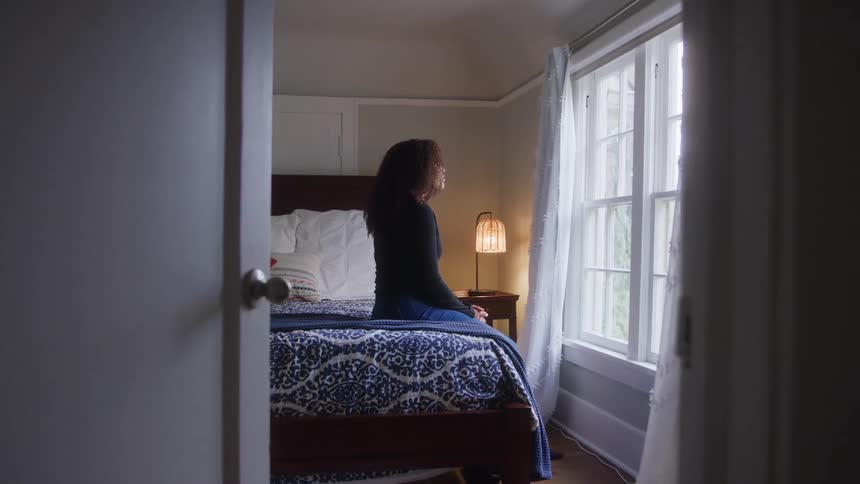A young woman sits on a bed looking forward with a pensive emotion bathed in natural light from a window
