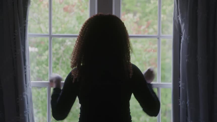 A woman with long curly hair is shown in silhouette from behind drawing open dark curtains to reveal a large window with a view of green foliage outsideafter fully opening the curtains she stands for a moment looking out the window illuminated by t
