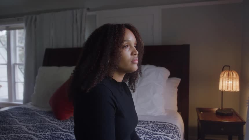 A depressed woman sits alone on a bed in a softly lit bedroom