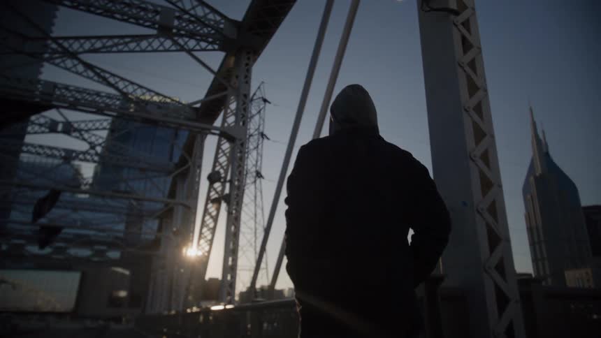 A figure in a dark hooded jacket is silhouetted on a metal truss bridge during sunrise or sunset with a bright sun flare visible on the leftthe person is facing away from the camera and towards a distant city skyline which includes a distinct spire