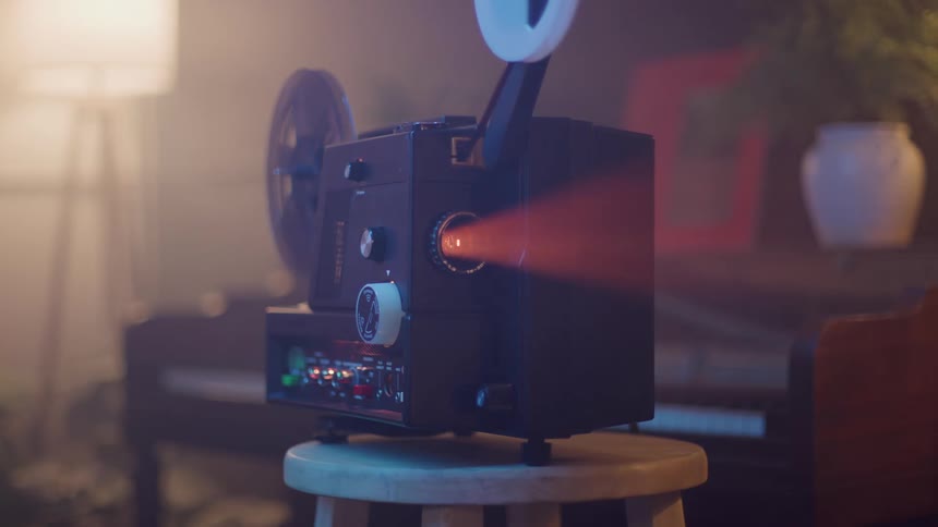 A vintagestyle film projector likely a super 8 or 16mm model is set up on a small wooden stool in a dimly lit roomthe machine is ready for operation with a film reel attached and the surrounding room is subtly illuminated by a floor lamp and featur