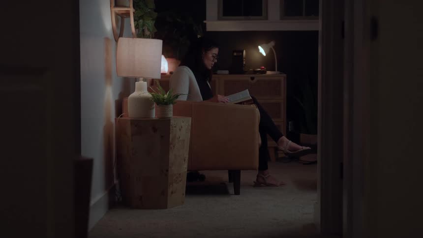 A woman sits in a chair reading a book by the light of a small table lamp at night