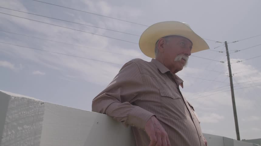 Older person in white hat leaning on wooden fence under partly cloudy sky