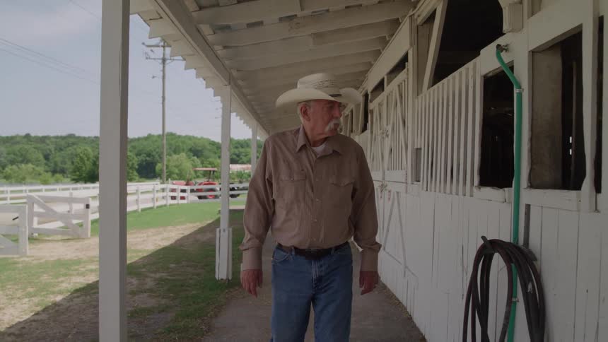 An older man walks through the shade of a white barn