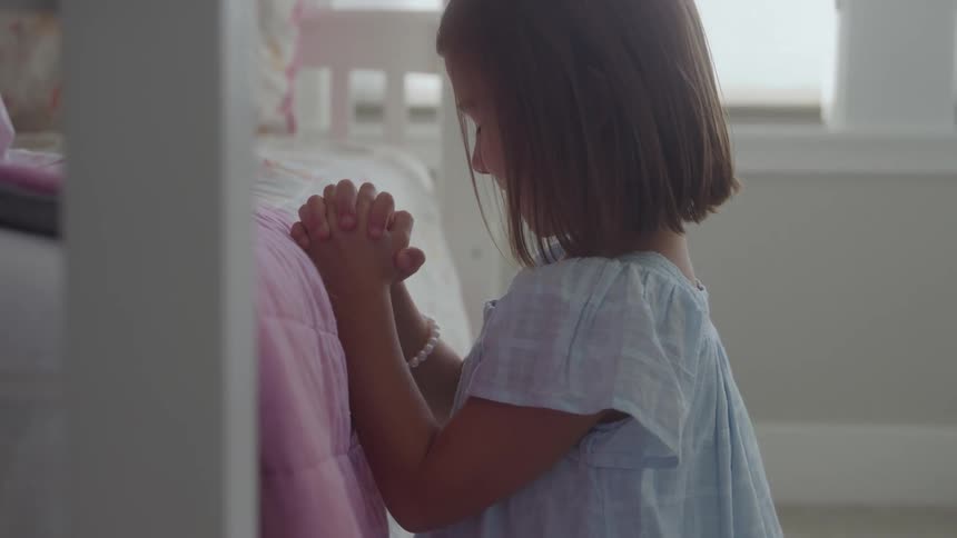 A young girl with shoulderlength brown hair kneeling beside a bed with a pink covering holding her hands tightly clasped together and bowed in prayer