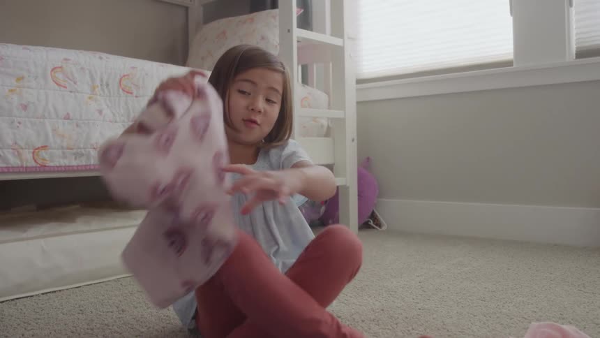 This short video shows a young girl sitting on a carpeted floor folding laundry in what appears to be a brightly lit bedroom with a bunk bed she is holding up a pink patterned piece of clothing possibly pajamas