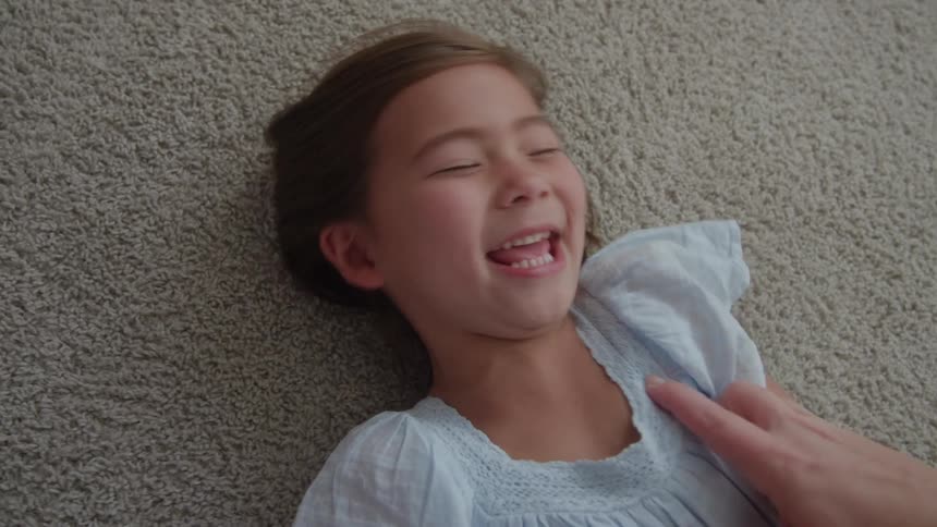 A young girl with brown hair is lying on a carpeted floor laughing joyfully a hand is visible near her seemingly tickling her arm which is causing her amusement
