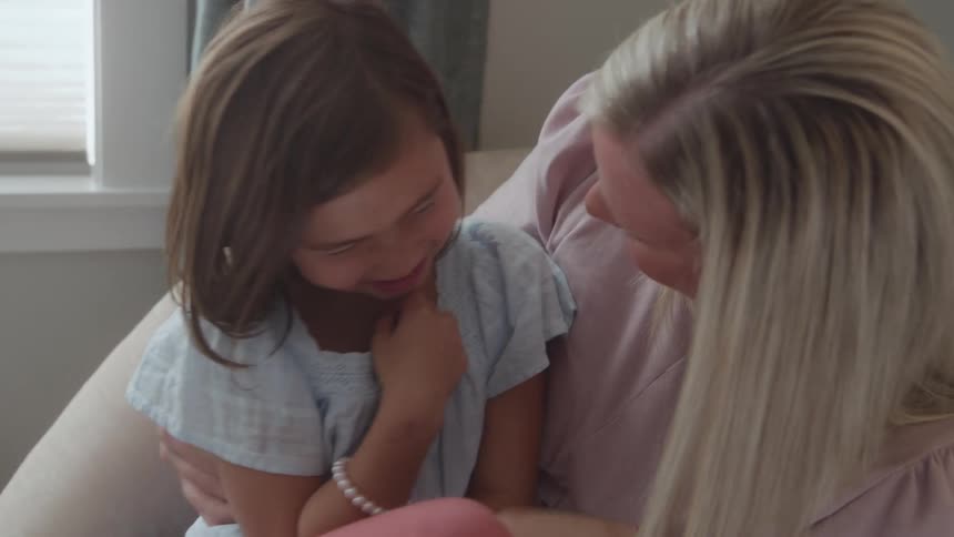 A smiling mother with blonde hair holds and embraces her young daughter with long brown hair sitting together in a bright soft lit room