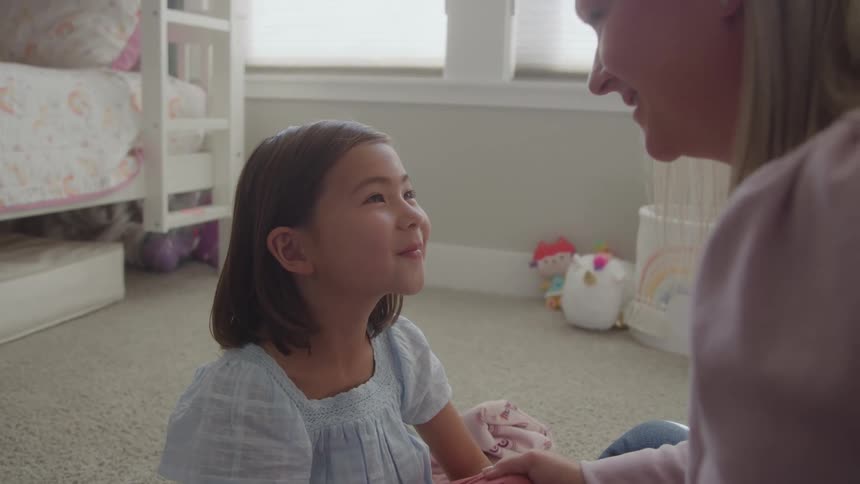 A young girl with dark hair wearing a light blue dress is sitting on the floor of what appears to be a childs bedroomshe is looking up and smiling warmly at a woman likely her mother who is sitting opposite her creating a sweet moment of connect