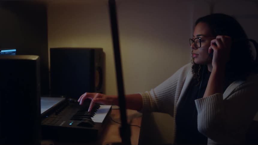 In the video a woman with glasses and headphones is sitting in a dimly lit studio space focused on playing a midi keyboard the scene suggests she is working on music production or composition late at night