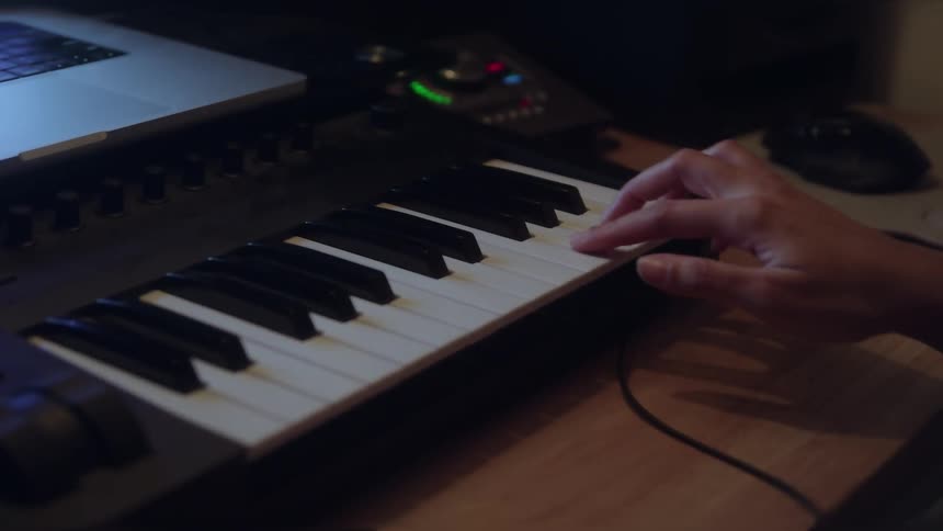 Person playing midi keyboard connected to laptop on wooden desk with lit audio interface in background