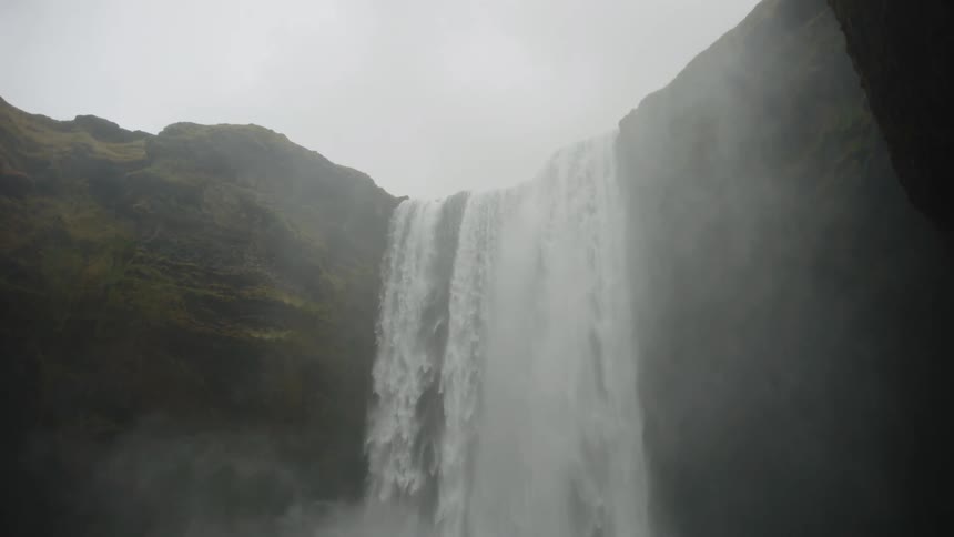 This video features a powerful wide waterfall plunging down a sheer dark cliff face under an overcast or misty sky the water creates a significant amount of spray and mist at the bottom