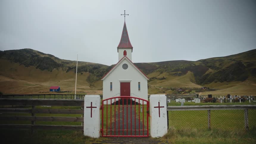 A small white church with a red roof stands in a green cloudy landscape beside a cemetery