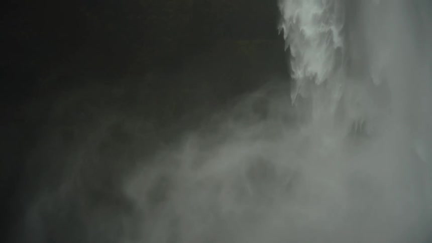 A closeup shot focuses on the immense power of a waterfall where a continuous column of white water cascades down the surrounding air is thick with mist and spray creating a high contrast against the dark background