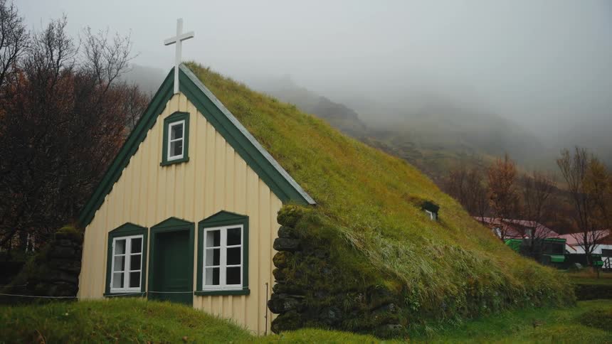 A yellow and green church with a turf roof and a cross stands against a foggy grassy hillside under a pale sky