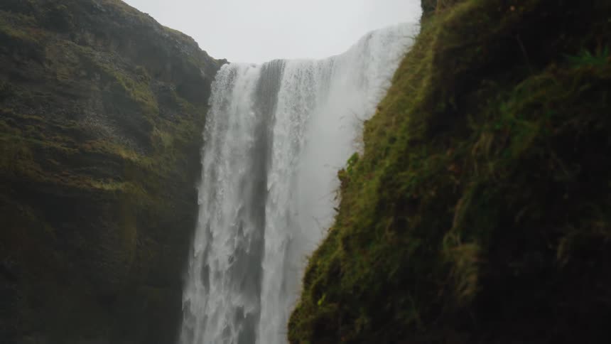 A large powerful waterfall cascades over a steep mossy cliff under an overcast sky
