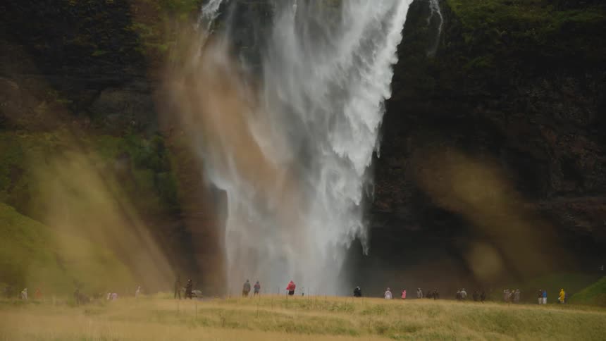 The video shows a powerful tall waterfall cascading down a dark cliff face surrounded by green slopesin the foreground a group of people appearing small in comparison to the falls are scattered across a wide grassy goldencolored field observing