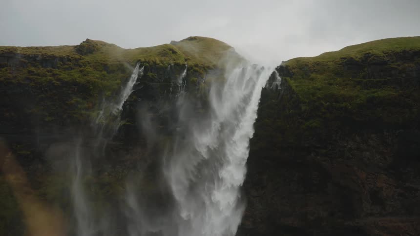 This video features a powerful multistream waterfall cascading down a sheer cliff face covered in lush green moss or grass at the top the scene is shot on an overcast day and the mist from the rushing water adds to the dramatic atmosphere