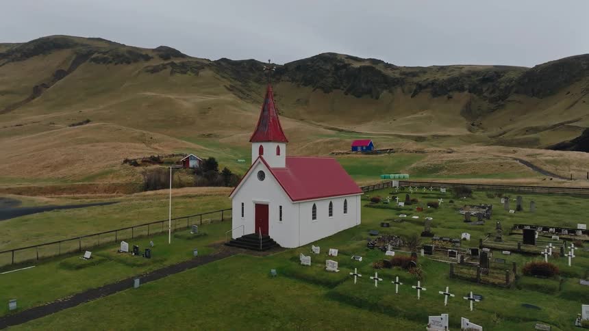 A small traditional white church with a distinctive red roof and spire stands in a grassy graveyardthe church and cemetery are set against a backdrop of rolling goldenbrown hills under an overcast sky suggesting a remote or rural location