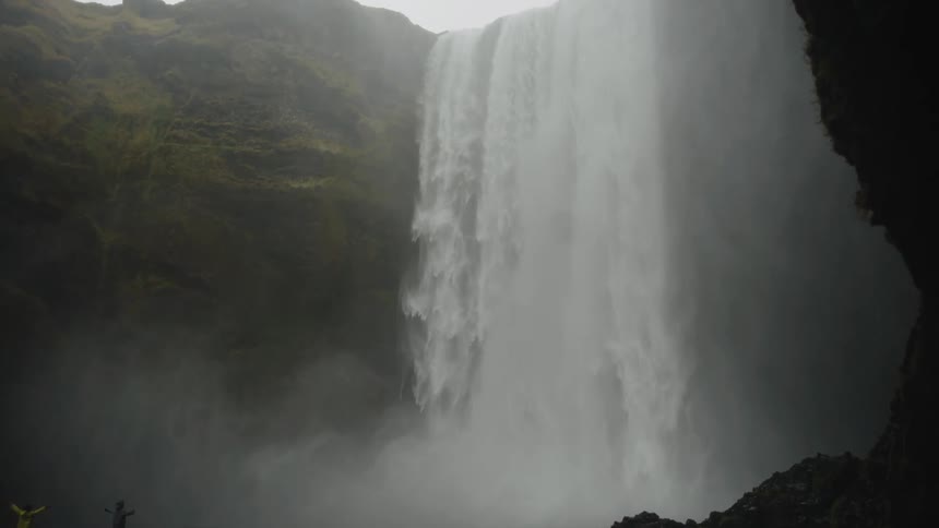 A person stands with arms out at the base of a massive waterfall
