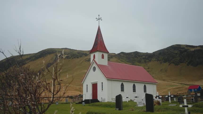 The video features a static shot of a quaint white wooden church with a striking red roof and tall steeple likely in a remote northern european settingthe church is situated on a grassy hill overlooking a small cemetery with large dramatic grassy