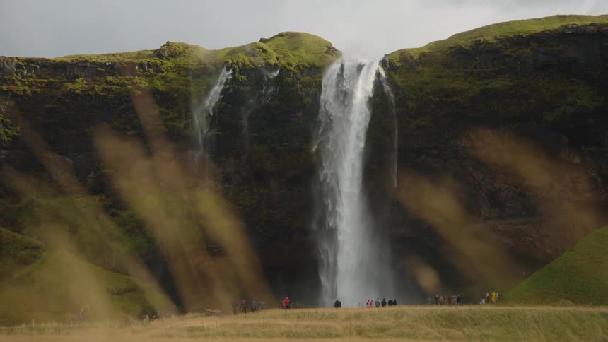 This video shows a powerful waterfall cascading over a mosscovered cliff face into a grassy field belowa small group of tourists is visible at the base of the falls dwarfed by the landscape with rays of sunlight occasionally cutting through the mis